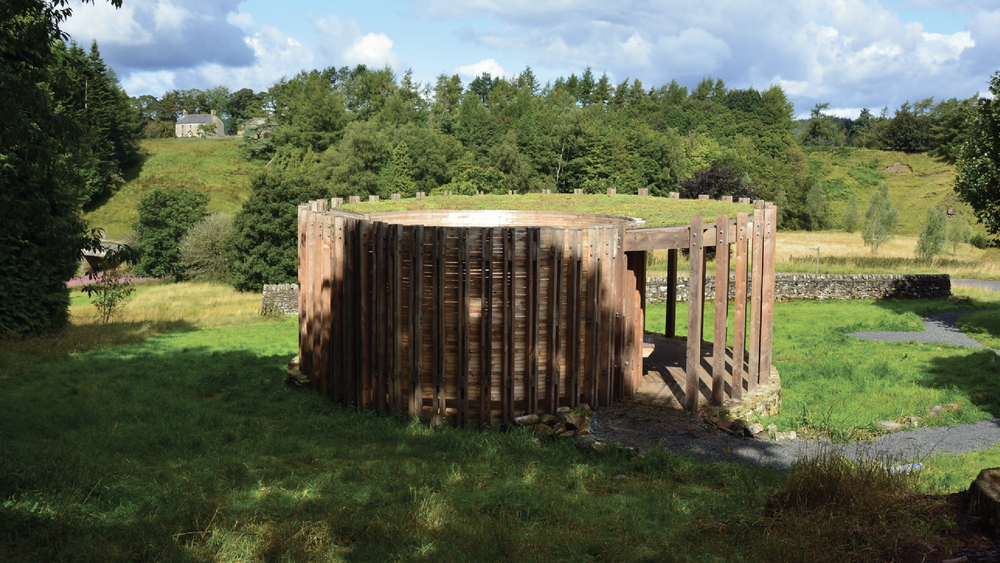 Wooden structure in a grassy field with trees and buildings in the background